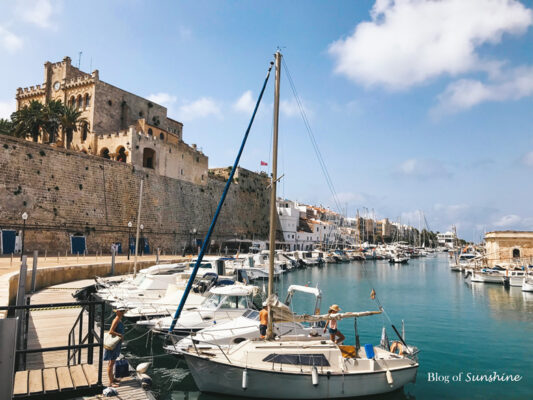 Boats moored at the Port of Ciutadella, Menorca with historic buildings along the marina under a bright blue sky