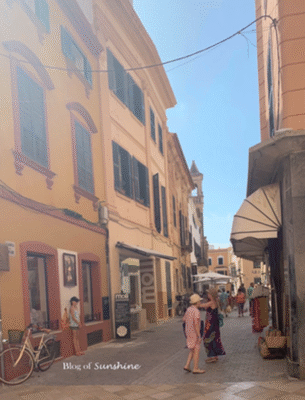 Sunlit side street in Ciutadella, Menorca with boutique shops, pedestrians, and colorful old town buildings