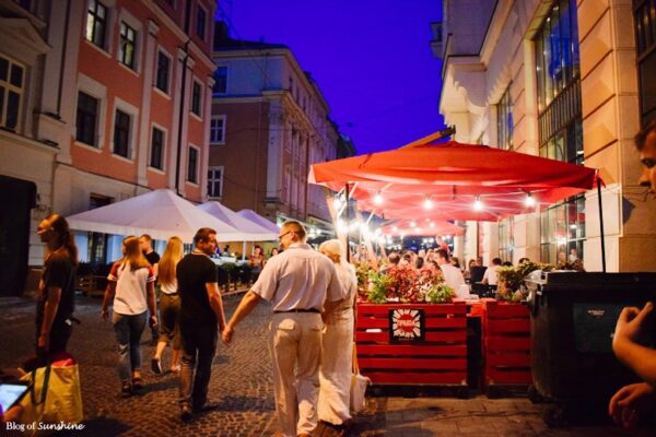 People walking past a lively café under red umbrellas on a cobbled street in Lviv during the evening