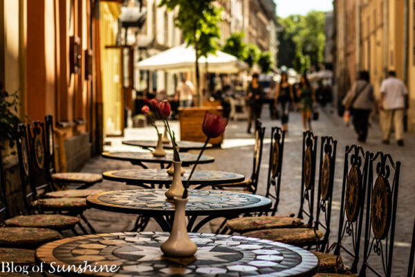 Empty mosaic tables at a cozy outdoor café in Lviv’s Old Town, with blurred pedestrians and sunlit buildings in the background