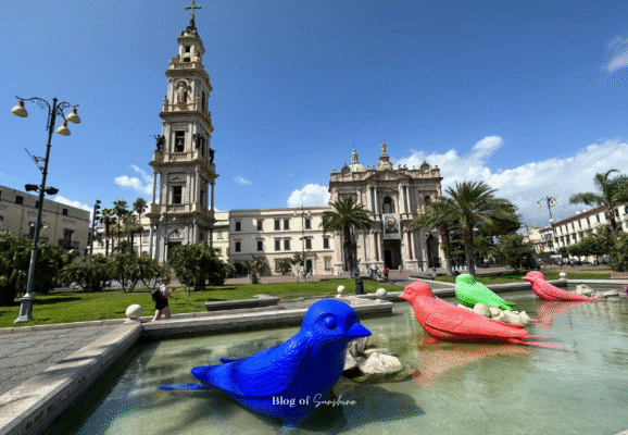 View of Piazza Bartolo Longo in modern Pompeii with colorful bird sculptures in a fountain and the Basilica of the Madonna of the Rosary in the background.