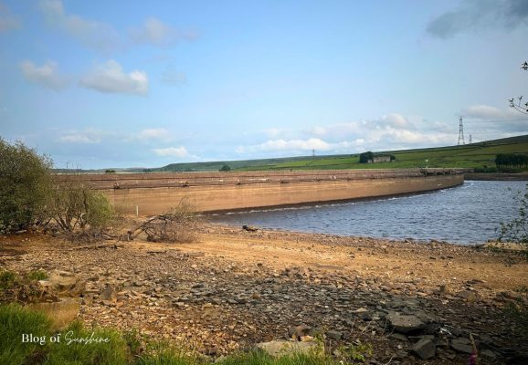 Wide view of Baitings Reservoir dam from the shoreline with exposed rocky foreshore and low water levels