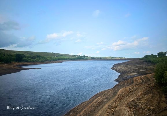 View from the bridge at Baitings Reservoir looking down the full length of the water with exposed shoreline and hills beyond