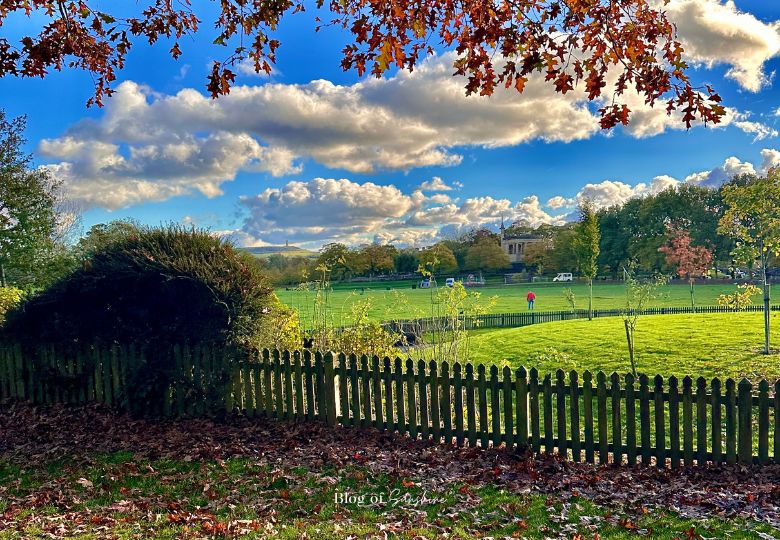 Autumn view across Greenhead Park Huddersfield with picket fence, lawns and trees under blue sky