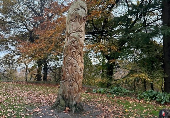 Tall wooden sculpture with intricate carvings of leaves and woodland wildlife surrounded by autumn trees at Beaumont Park in Huddersfield.