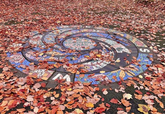 Circular mosaic at Beaumont Park in Huddersfield, with colourful tiled patterns partly covered by autumn leaves.