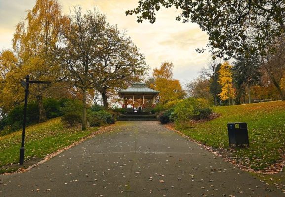 Paved walking path leading to the bandstand at Beaumont Park in Huddersfield, framed by autumn trees.