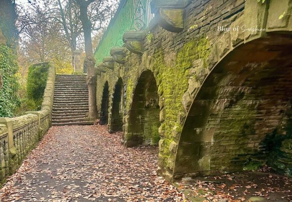 Moss-covered Victorian stone arches beside a leaf-covered pathway and stone steps at Beaumont Park in Huddersfield.